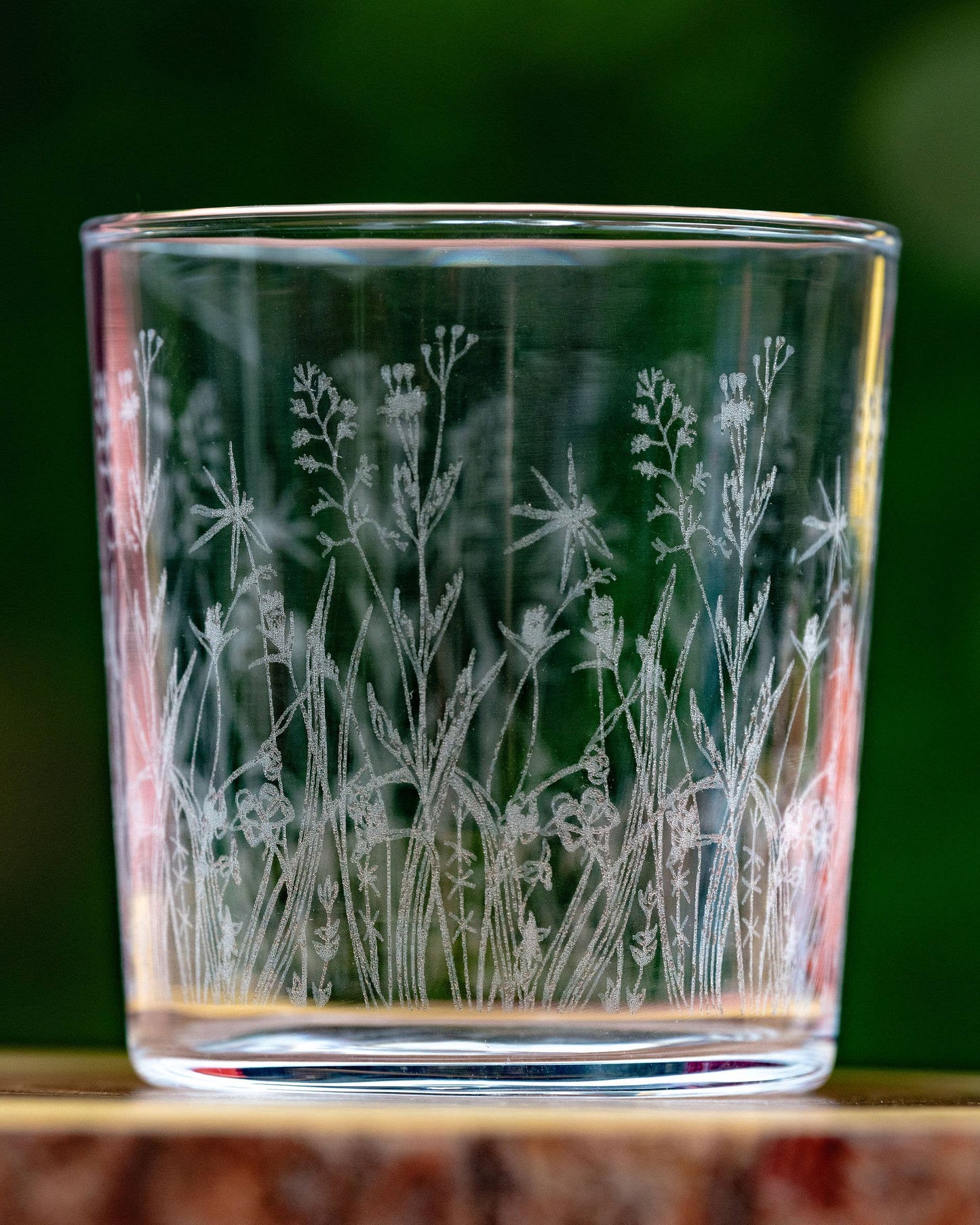 Glass tumbler with engraved floral design on a wooden surface with a blurred green background
