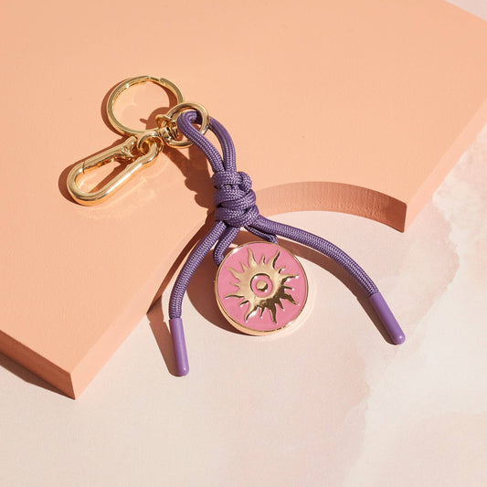 Bag charm and keychain with purple cord and pink disc featuring a sun design in gold