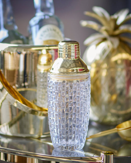 Decorative cocktail shaker with gold accents on a bar tray with bottles and a pineapple.