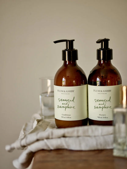 Two dark brown glass bottles, black pump dispensers, white labels with sage green writing, seaweed and samphire shampoo and conditioner by Plum and Ashby