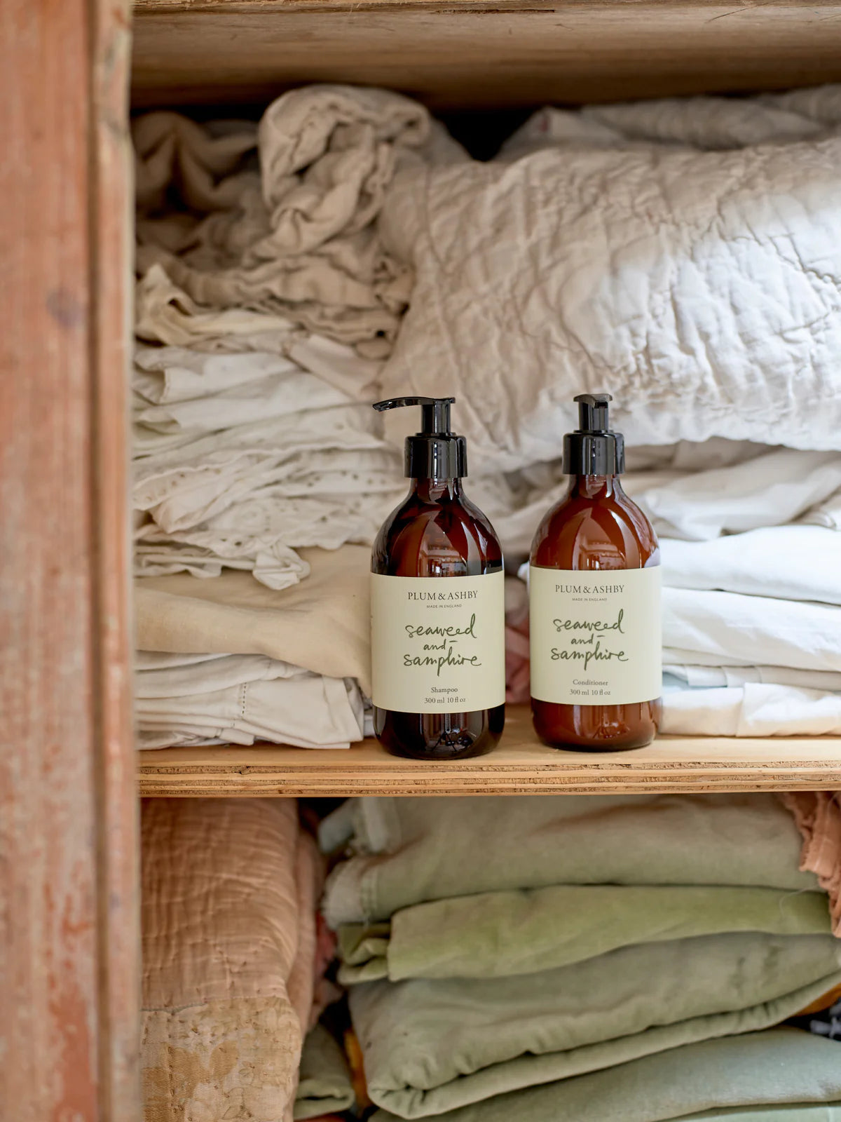 Laundry cupboard with two dark brown glass bottles, black pump dispensers, white labels with sage green writing, seaweed and samphire shampoo and conditioner by Plum and Ashby