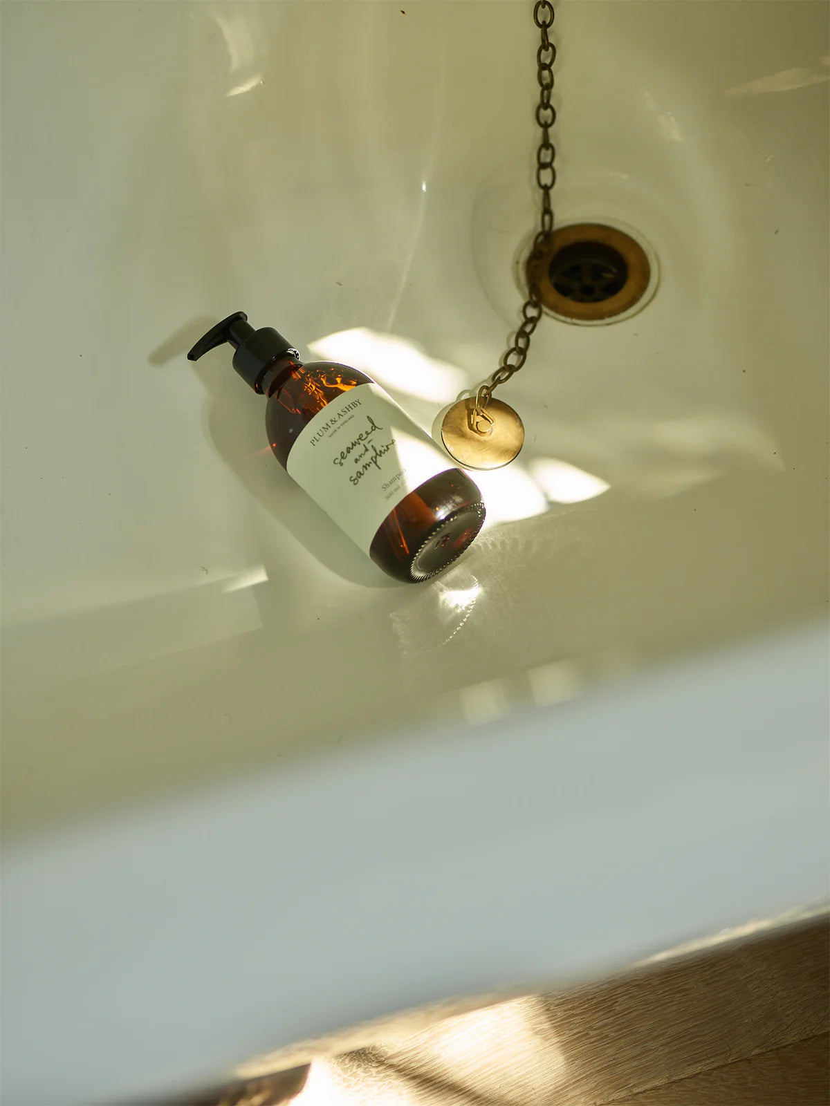 A bath and plug hole with a dark brown glass bottle, black pump dispensers, white label with sage green writing, seaweed and samphire shampoo by Plum and Ashby