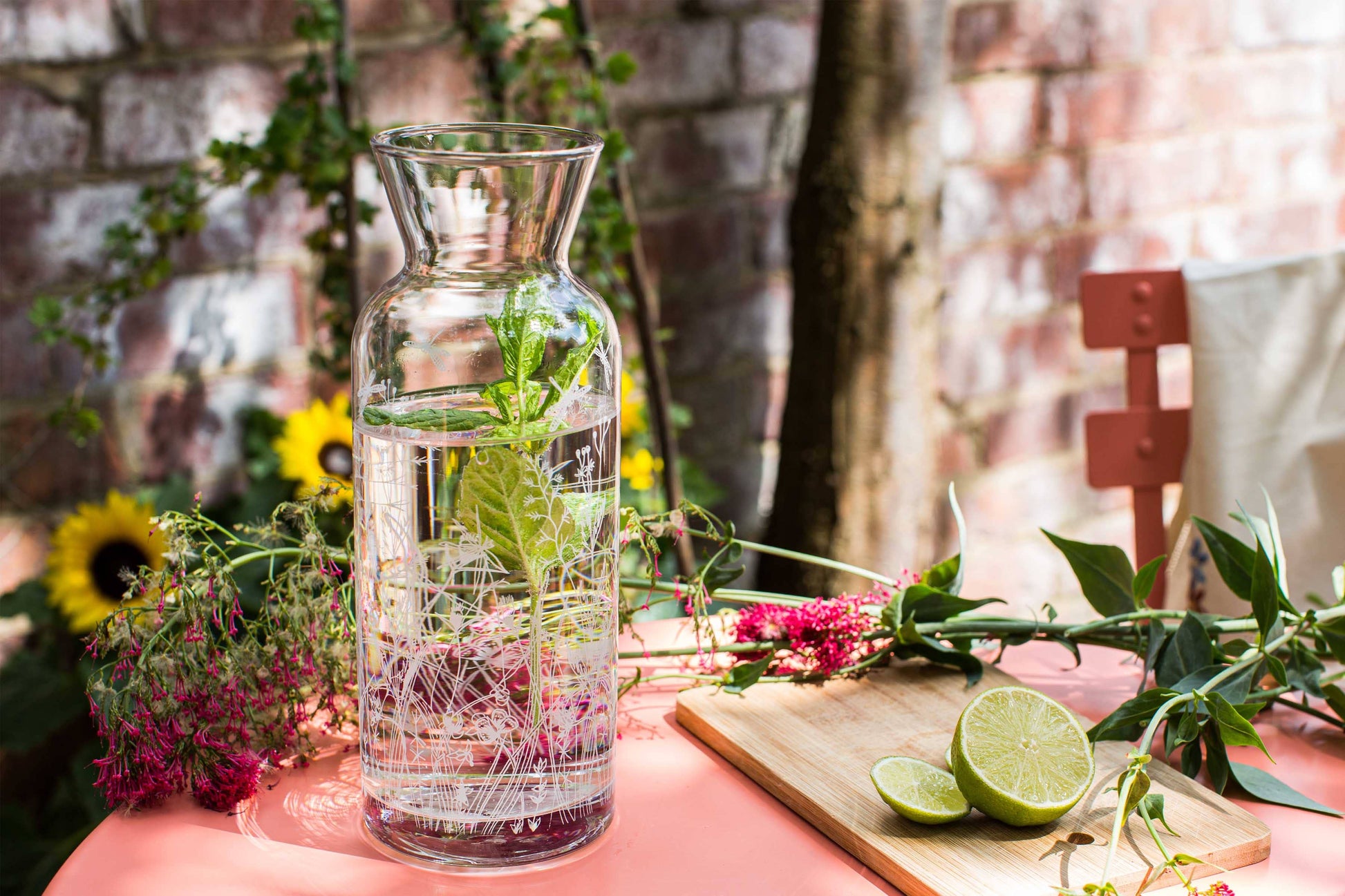 Glass carafe with water and herbs on a table with flowers and a cutting board in the background.