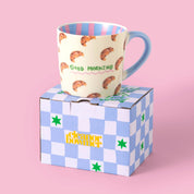 Mug with croissant pattern and 'Good Morning' text by Eleanor Bowmer, on top of blue and white check gift box.