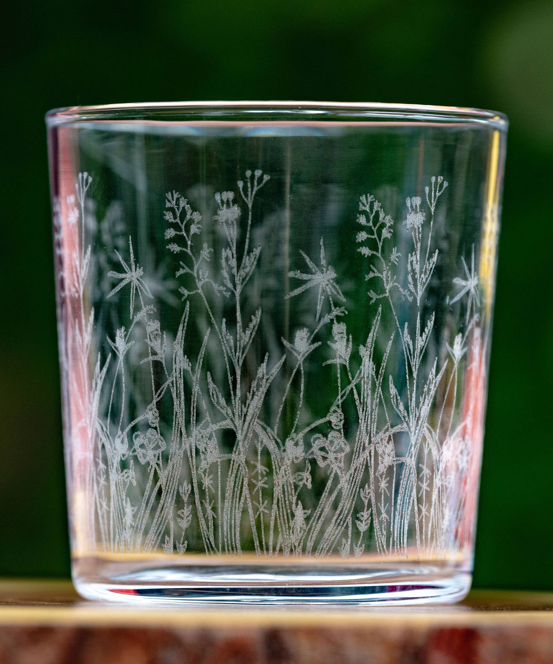 Glass tumbler with engraved floral design on a wooden surface with a blurred green background