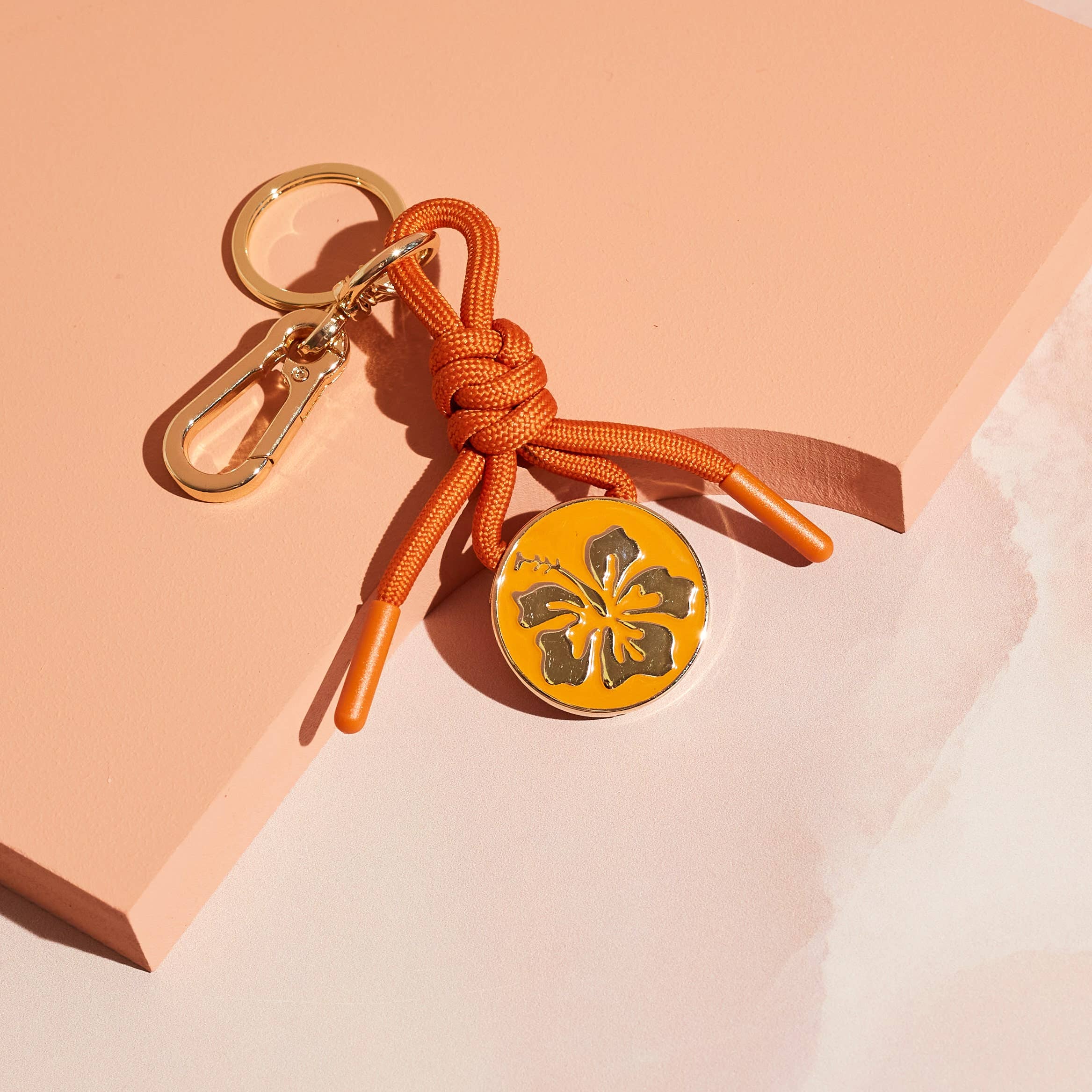 Bag charm keychain with orange cord and gold hibiscus emblem on a yellow charm