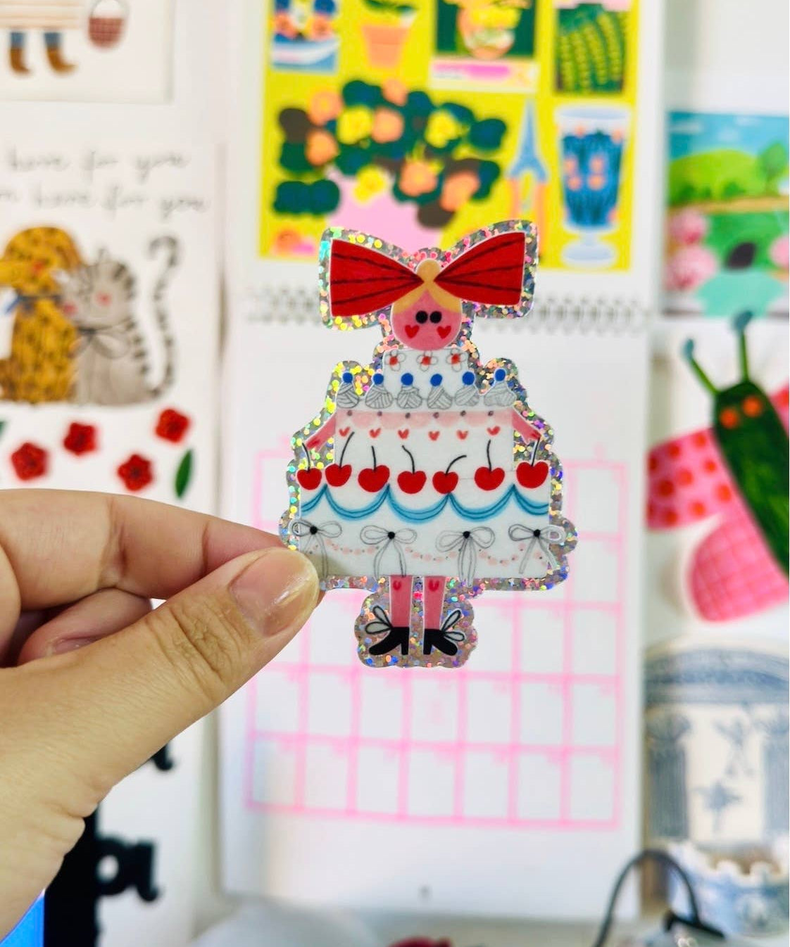 A colourful glitter sticker of a girl dessed as a birthday cake, held in front of a colourful wall with cartoon-style decorations.