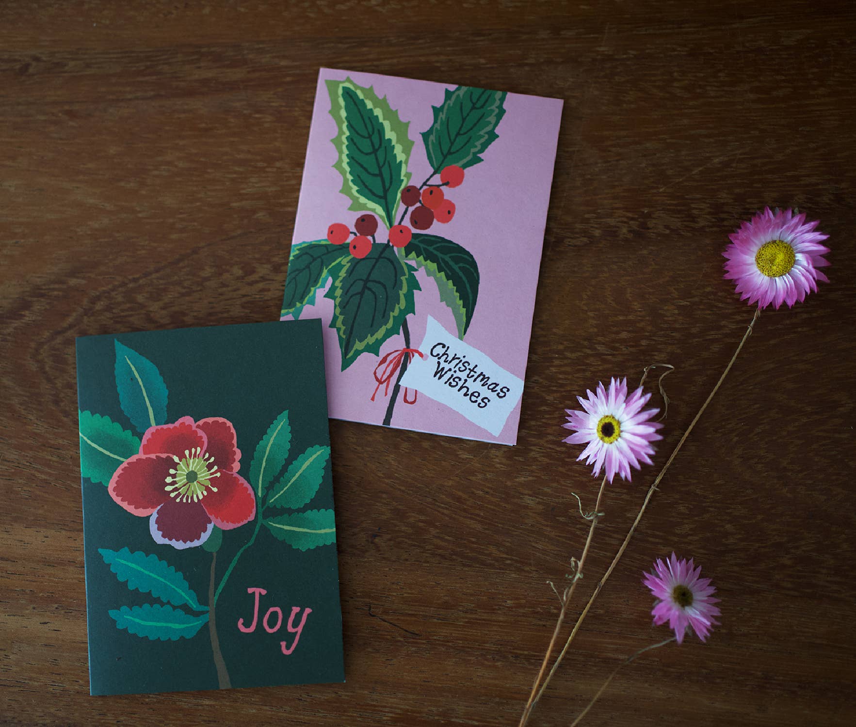 Two illustrated Christmas greeting cards arranged on a wooden surface, with dried pink flowers beside them. One card is dark green with a red flower and the word "Joy," and the other is light pink with green holly leaves, red berries, and a small "Christmas Wishes" note.