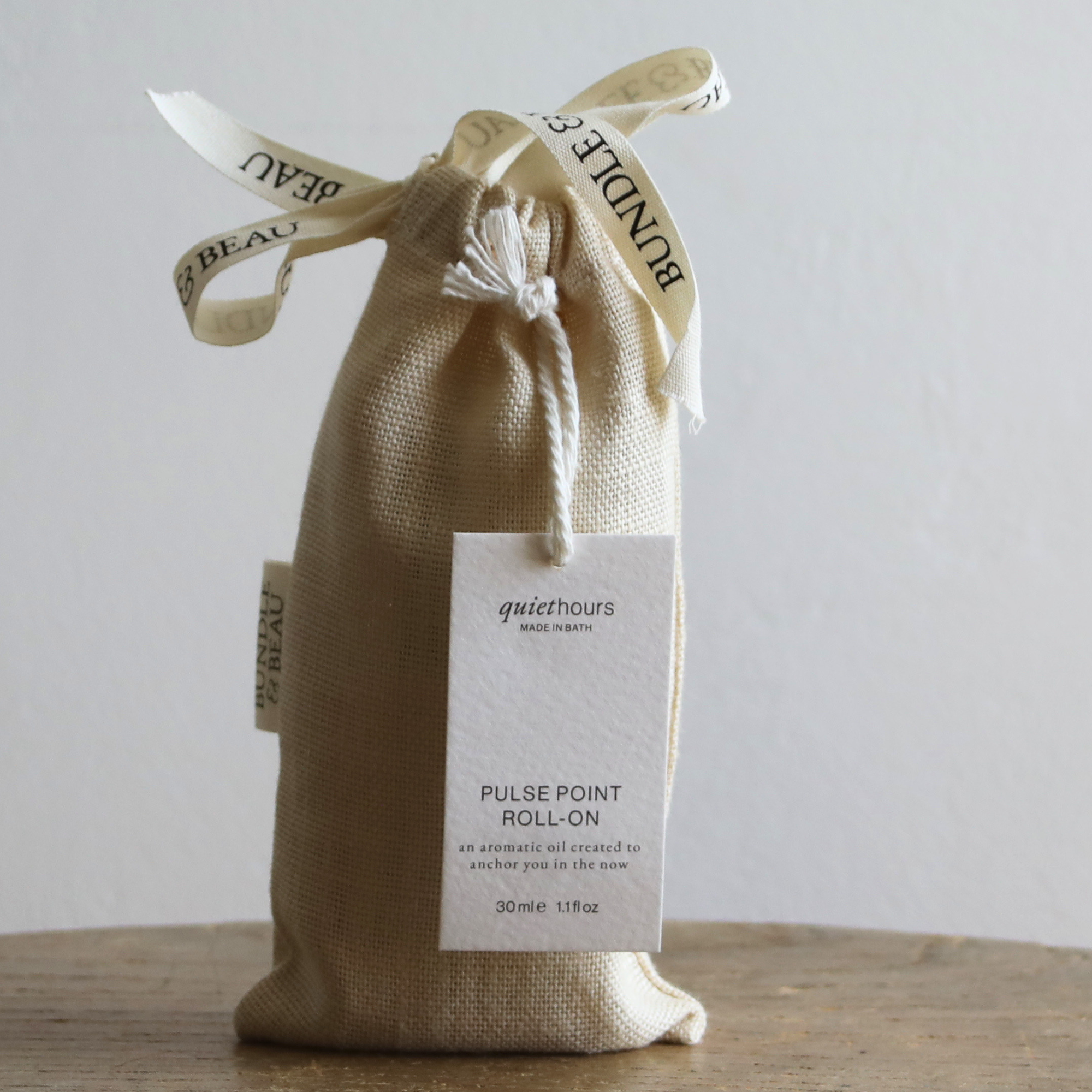 A small beige burlap drawstring pouch tied with a cream ribbon  holding a white product tag that reads 'Quiet Hours Pulse Point Roll-on.