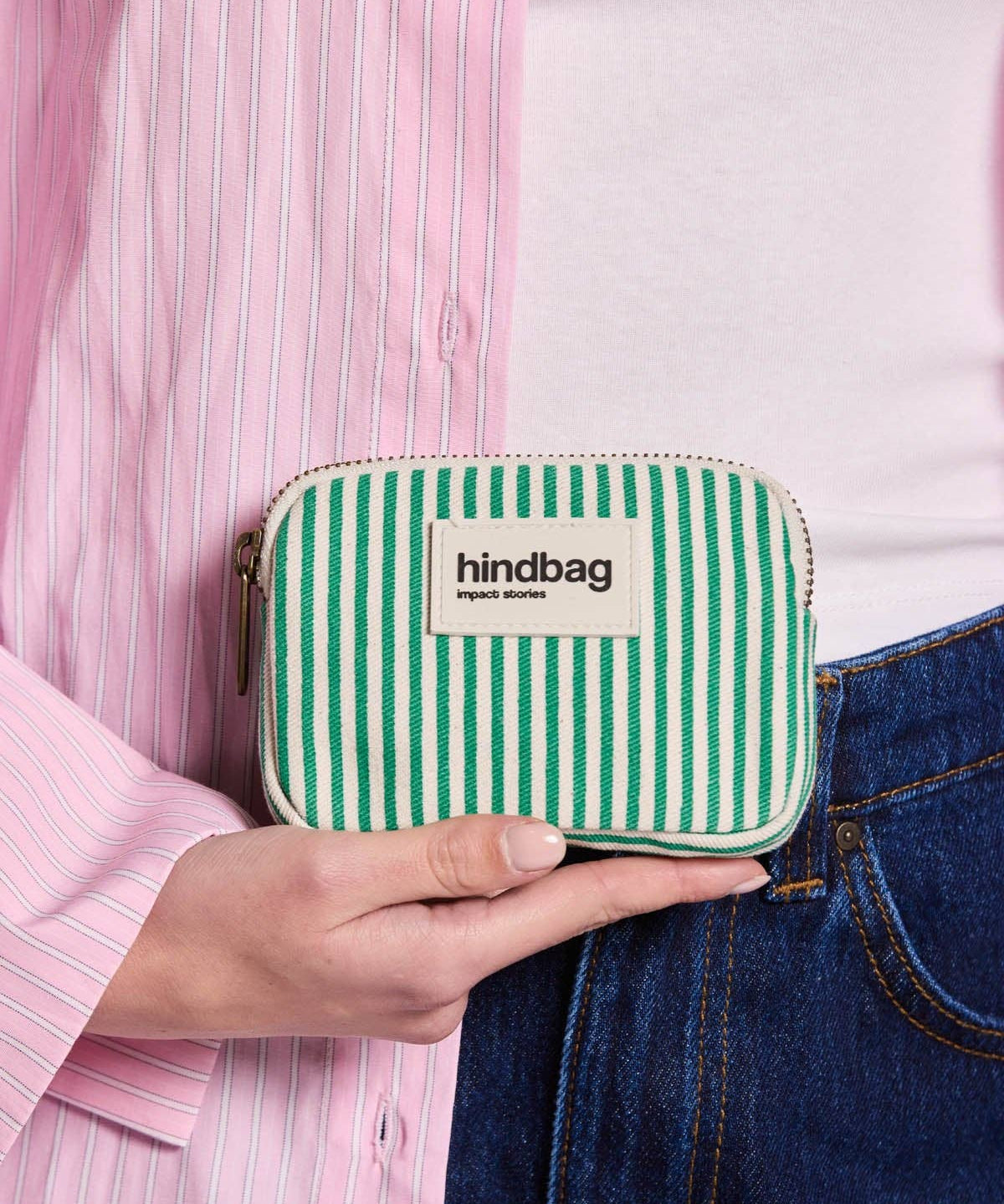 Green and white striped coin purse or pouch with 'hindbag' label held by a person wearing a pink shirt and blue jeans.