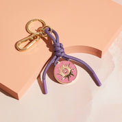 Bag charm and keychain with purple cord and pink disc featuring a sun design in gold