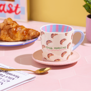 Mug with croissant pattern and 'Good Morning' text by Eleanor Bowmer on a pink table with croissant and newspaper.