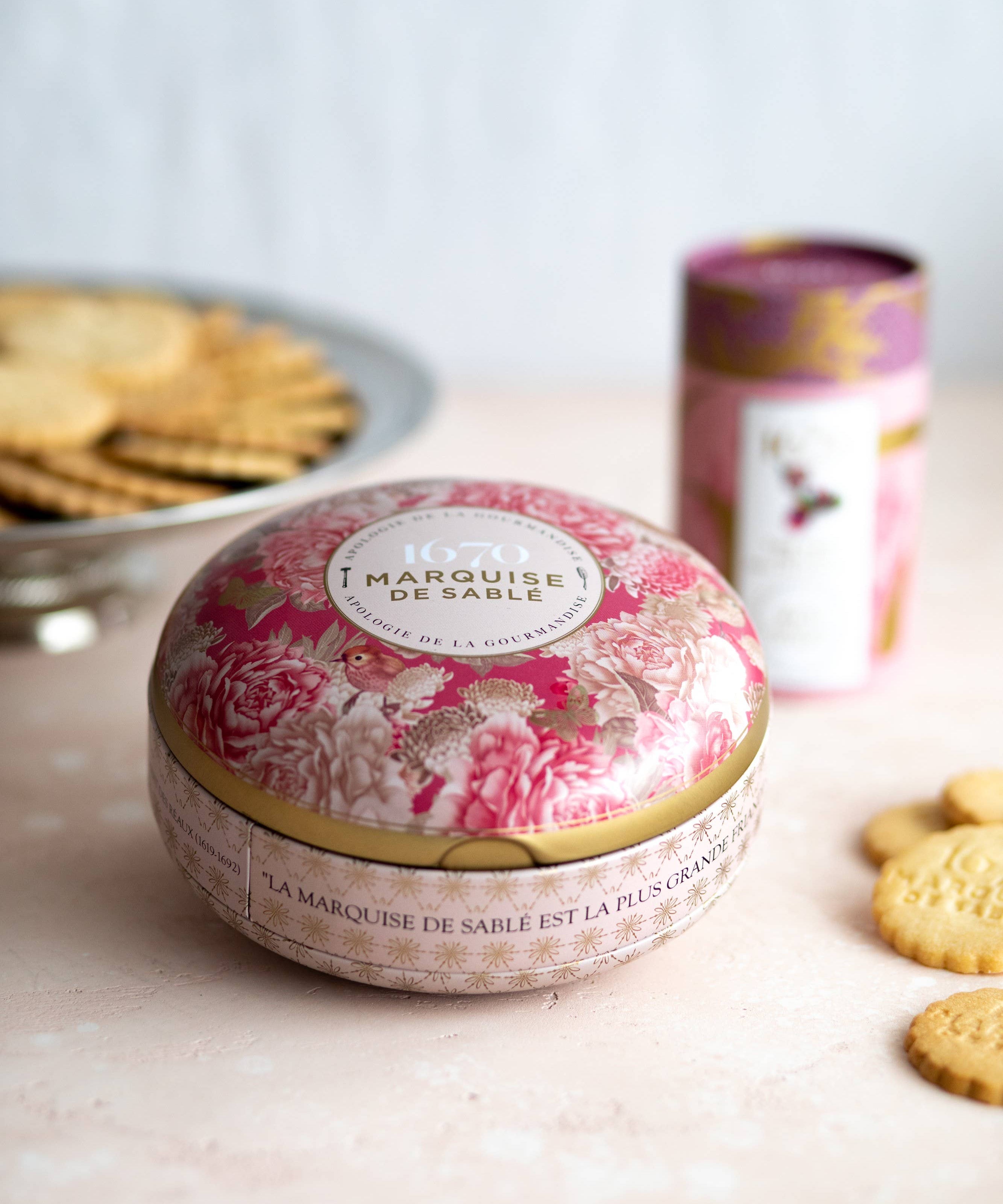 A round tin of Marquisse de Sable cookies in the foreground, decorated with a pink and beige floral pattern and gold trim, with other cookies and a matching tin blurred in the background on a light surface.