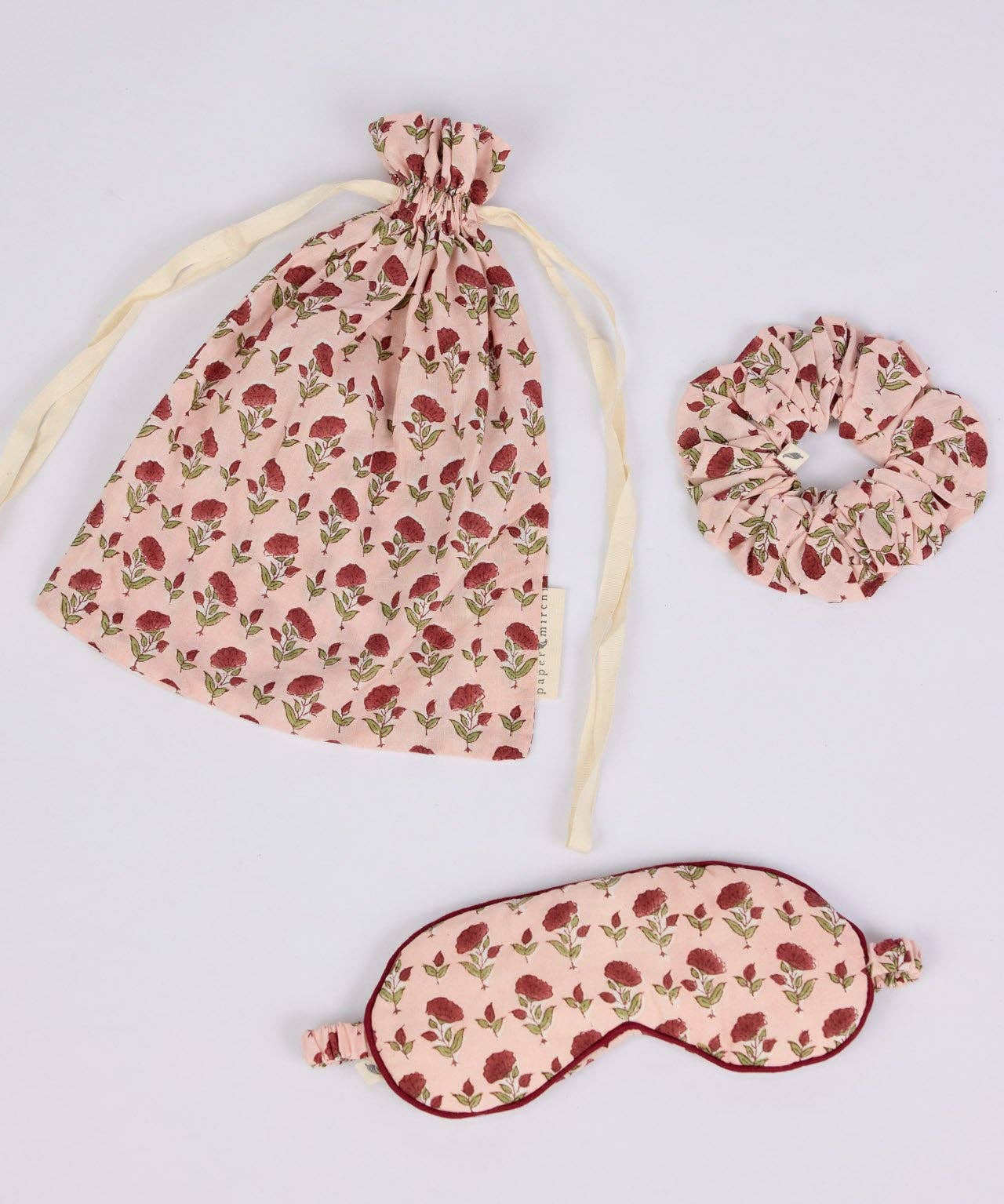 Three-piece sleep set featuring a drawstring bag, a scrunchie, and an eye mask in a dusty pink fabric with a red rose print, arranged on a light gray background.
