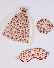 Three-piece sleep set featuring a drawstring bag, a scrunchie, and an eye mask in a dusty pink fabric with a red rose print, arranged on a light gray background.
