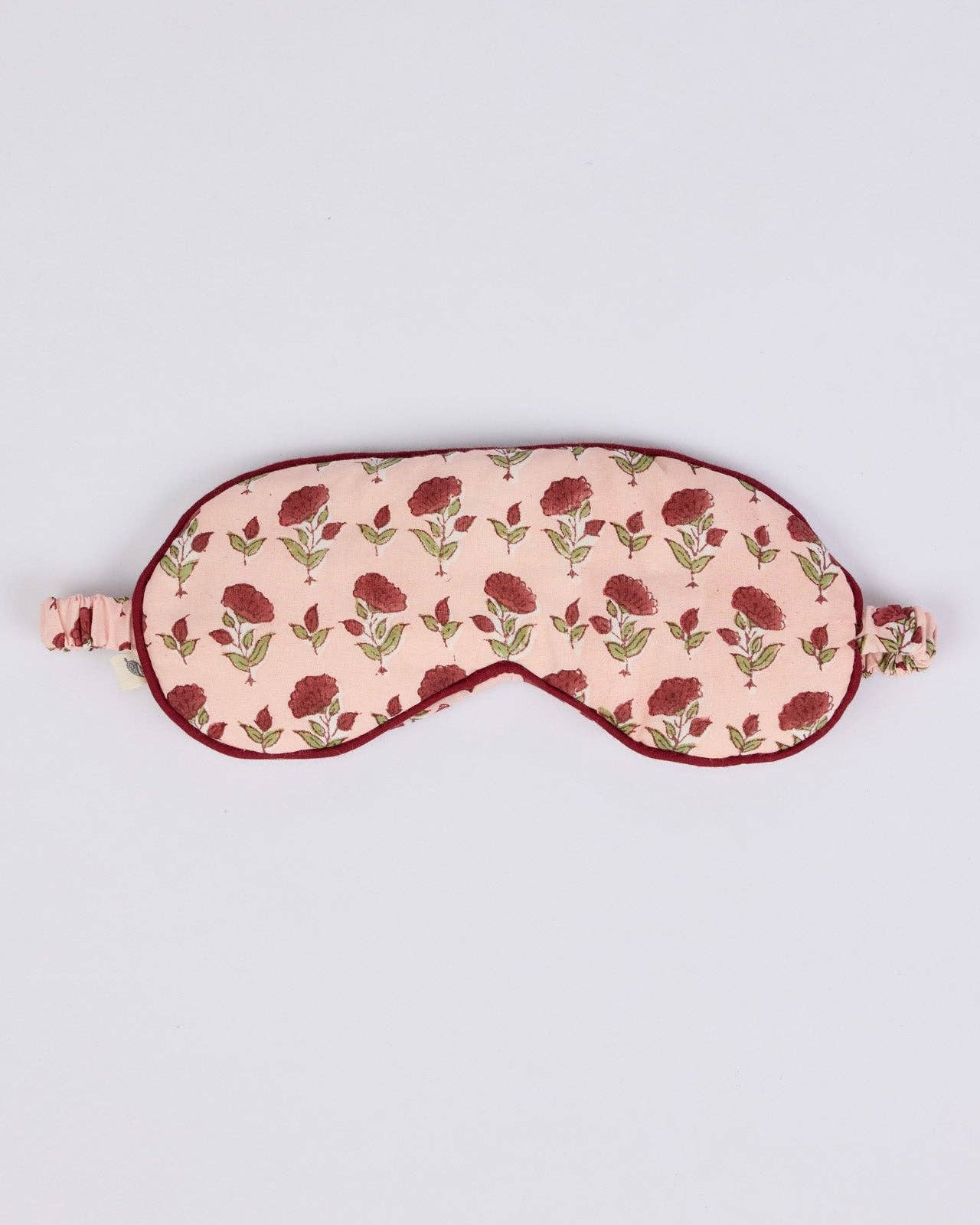 A fabric sleep mask with a light pink background, a repeating dark red rose pattern, and dark red trim, shown on a plain light gray background.