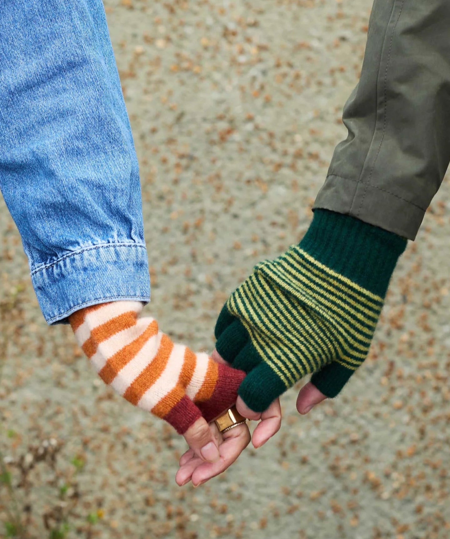 Two people holding hands while wearing fingerless knit gloves. One glove is orange and white striped with burgundy accents, and the other is dark green with yellow-green horizontal stripes.