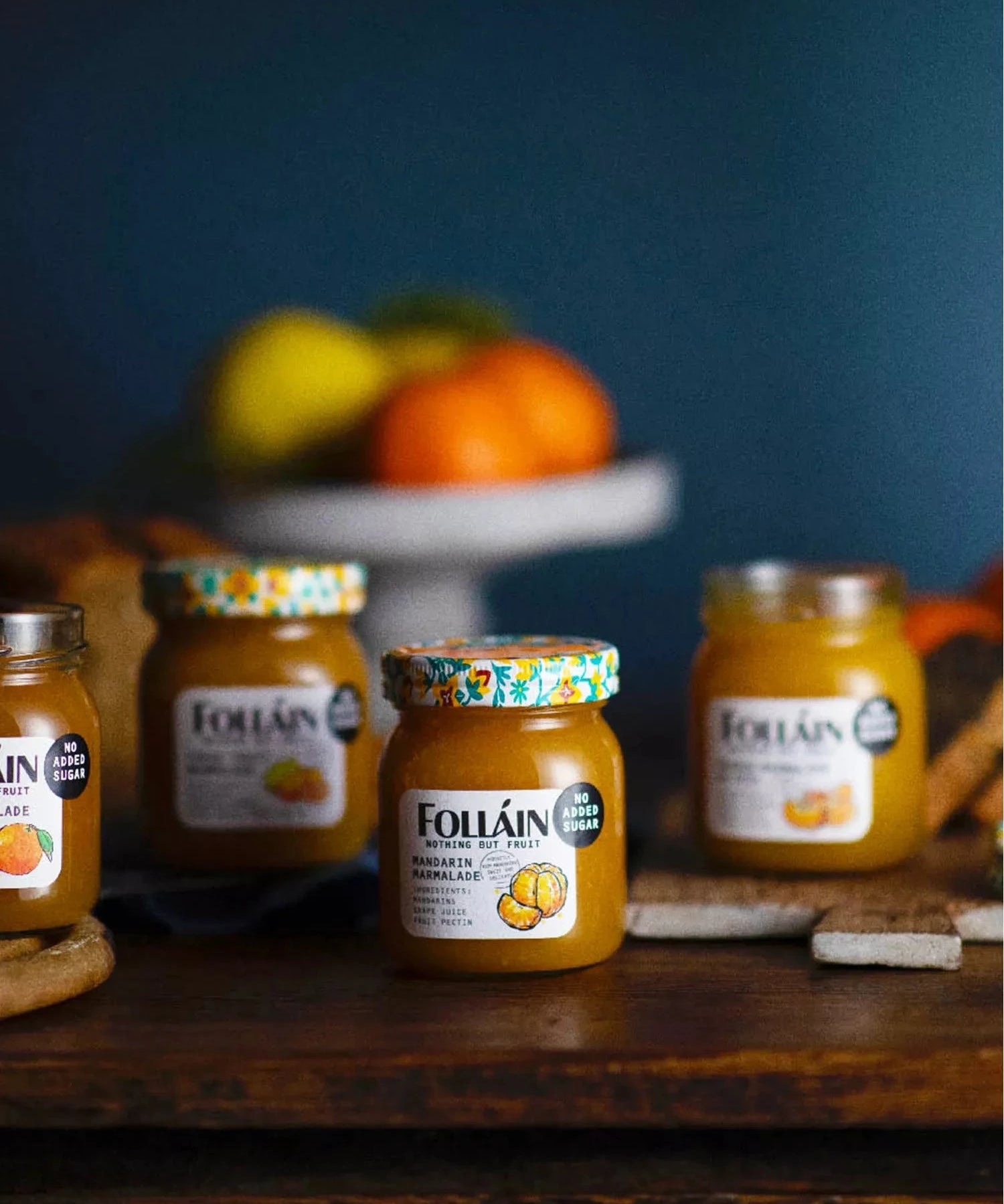 Four jars of Follain pumpkin and sage jam arranged on a dark wooden table with a blurred blue background and fruit on a stand behind them.