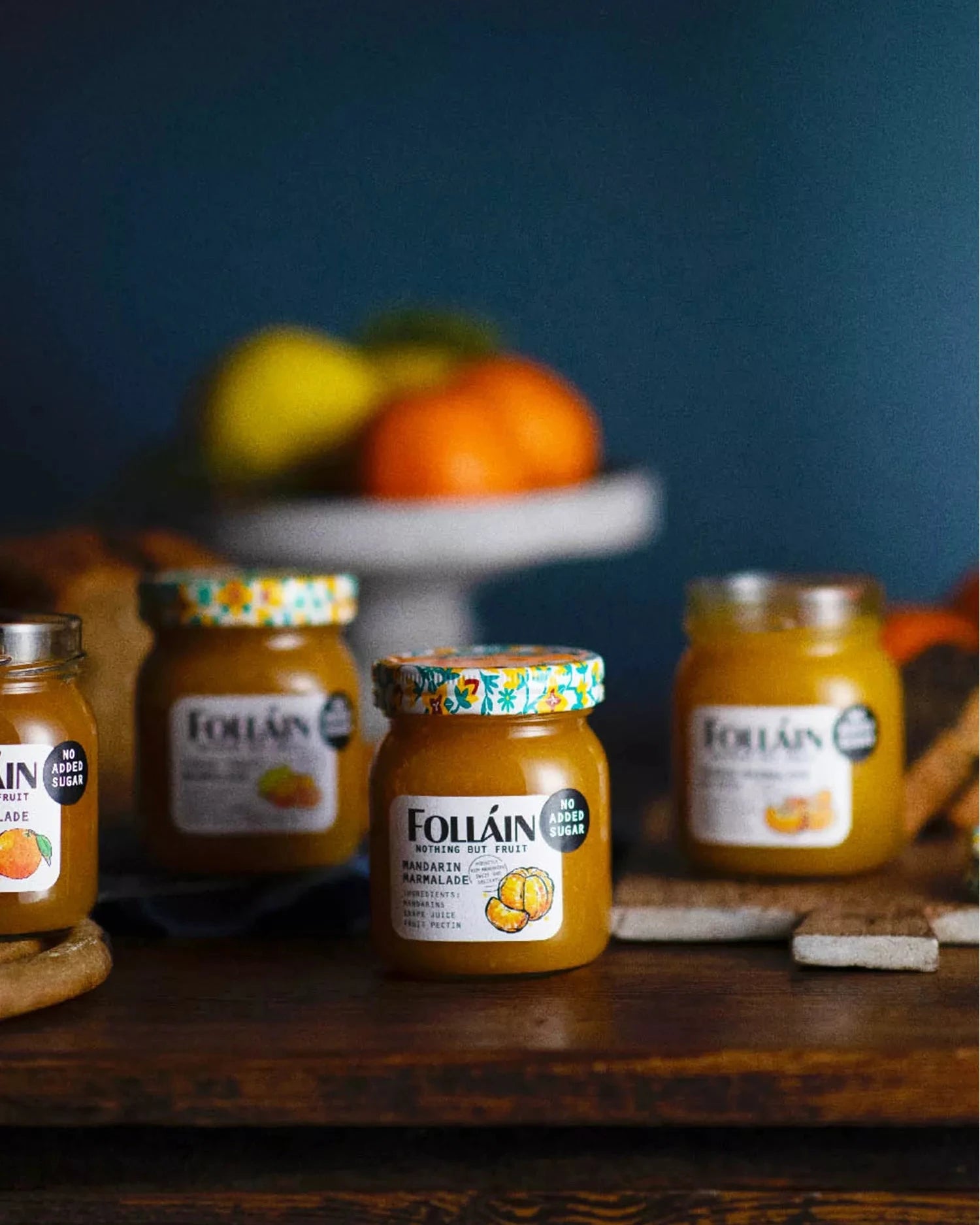 Four jars of Follain pumpkin and sage jam arranged on a dark wooden table with a blurred blue background and fruit on a stand behind them.