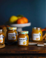Four jars of Follain pumpkin and sage jam arranged on a dark wooden table with a blurred blue background and fruit on a stand behind them.
