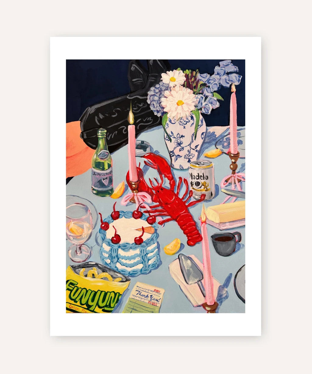 A framed art print featuring a colorful still life illustration of a lobster, cake, flowers, candles, and a bottle on a table, shown against a light beige background.