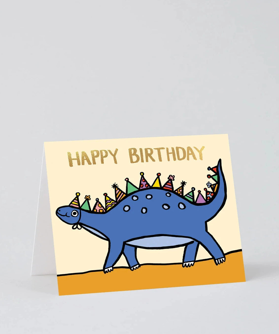 A square birthday card with a blue dinosaur wearing colorful party hats, standing on a gold base against a light gray background. The card reads 'HAPPY BIRTHDAY' in gold lettering on a cream background.