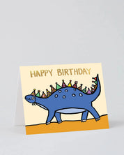 A square birthday card with a blue dinosaur wearing colorful party hats, standing on a gold base against a light gray background. The card reads 'HAPPY BIRTHDAY' in gold lettering on a cream background.