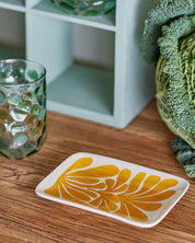 Decorative coaster with yellow floral design on a wooden surface next to a glass and plant.