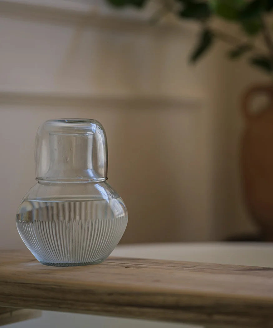 A small clear ribbed glass vase or candle holder sits on a light wooden table in a softly lit interior, with a blurred potted plant in the background.