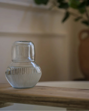 A small clear ribbed glass vase or candle holder sits on a light wooden table in a softly lit interior, with a blurred potted plant in the background.