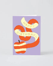 A square birthday greeting card with a lavender background, featuring a stylized orange and cream striped apple and ribbon banner that reads 'HAPPY BIRTHDAY!!' in cream lettering. The card is photographed standing upright on a light gray background.