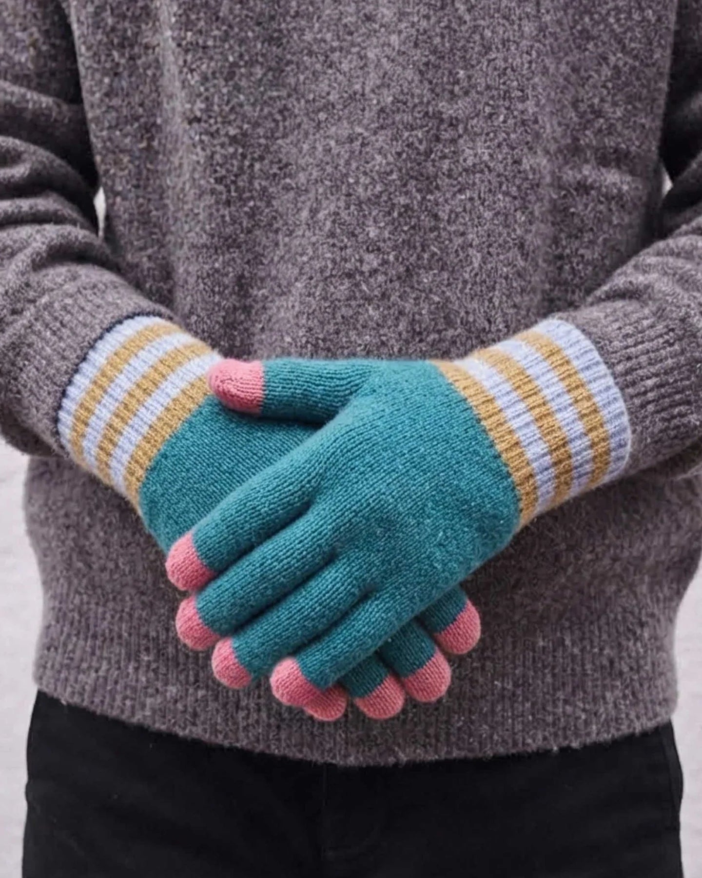A kid wearing teal knit gloves with pink fingertips and striped cuffs in yellow, white, and gray, shown against a light gray background.