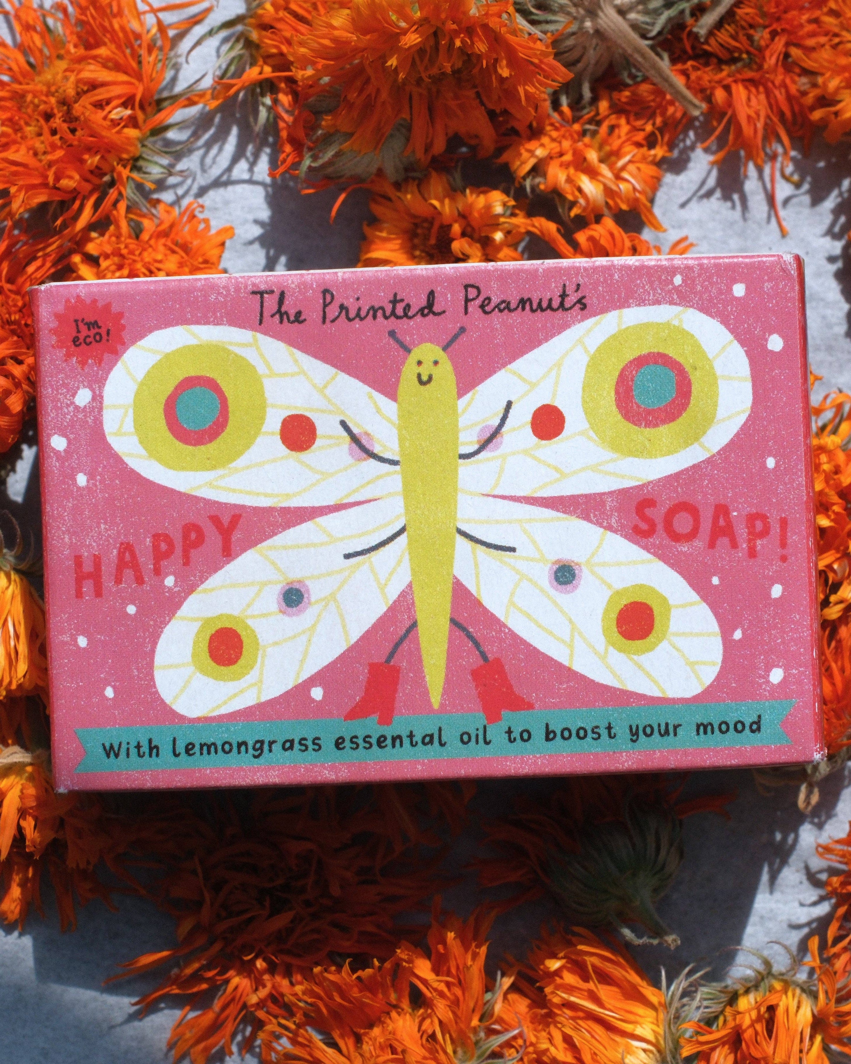 A rectangular pink soap bar box from 'The Printed Peanut's Happy Soap!' featuring a whimsical yellow and white butterfly illustration, styled on a gray surface surrounded by dried orange marigold flowers.