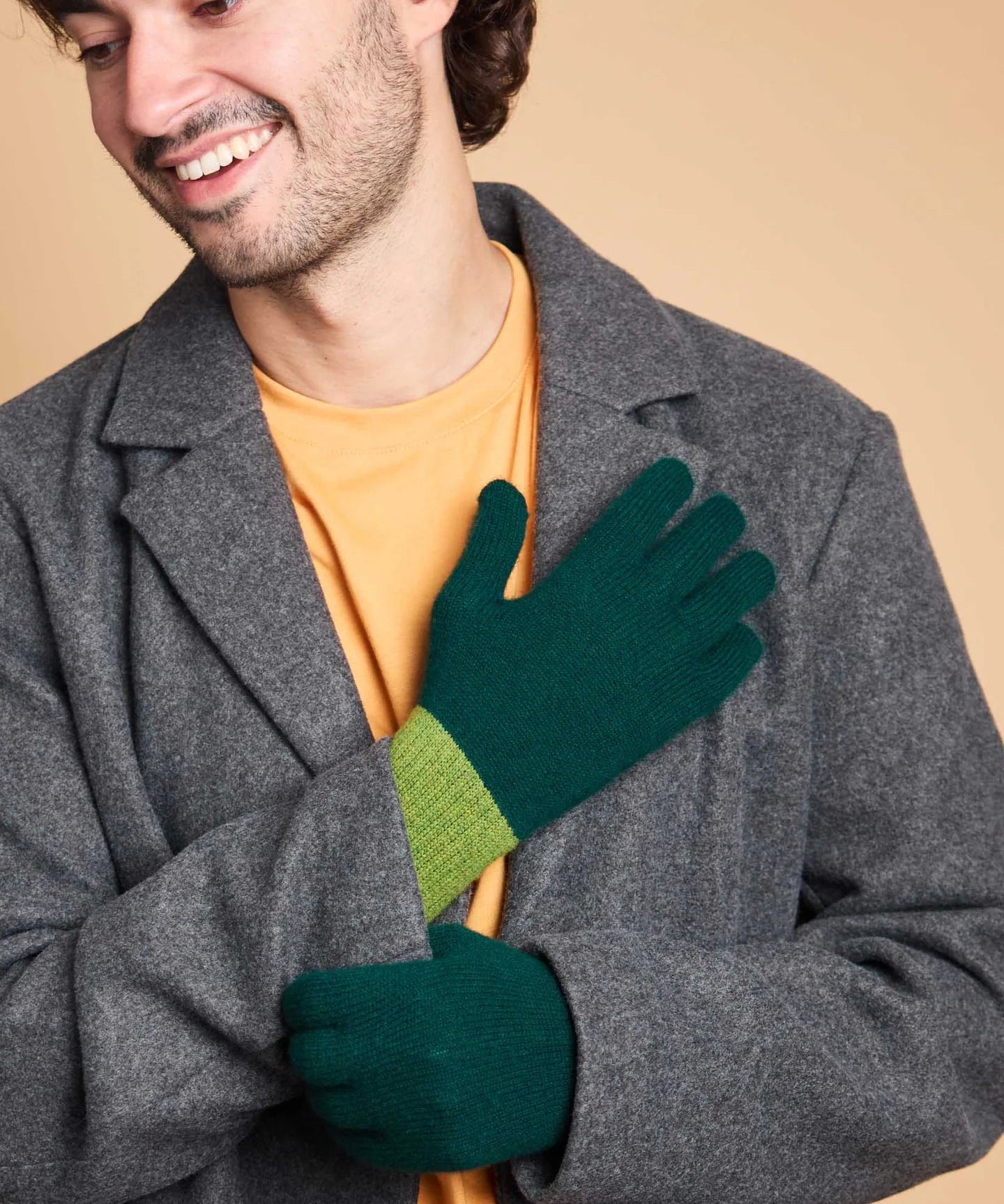 A man wearing a gray wool coat over a yellow-orange t-shirt, showcasing a pair of dark green knit gloves with a lime green ribbed cuff.