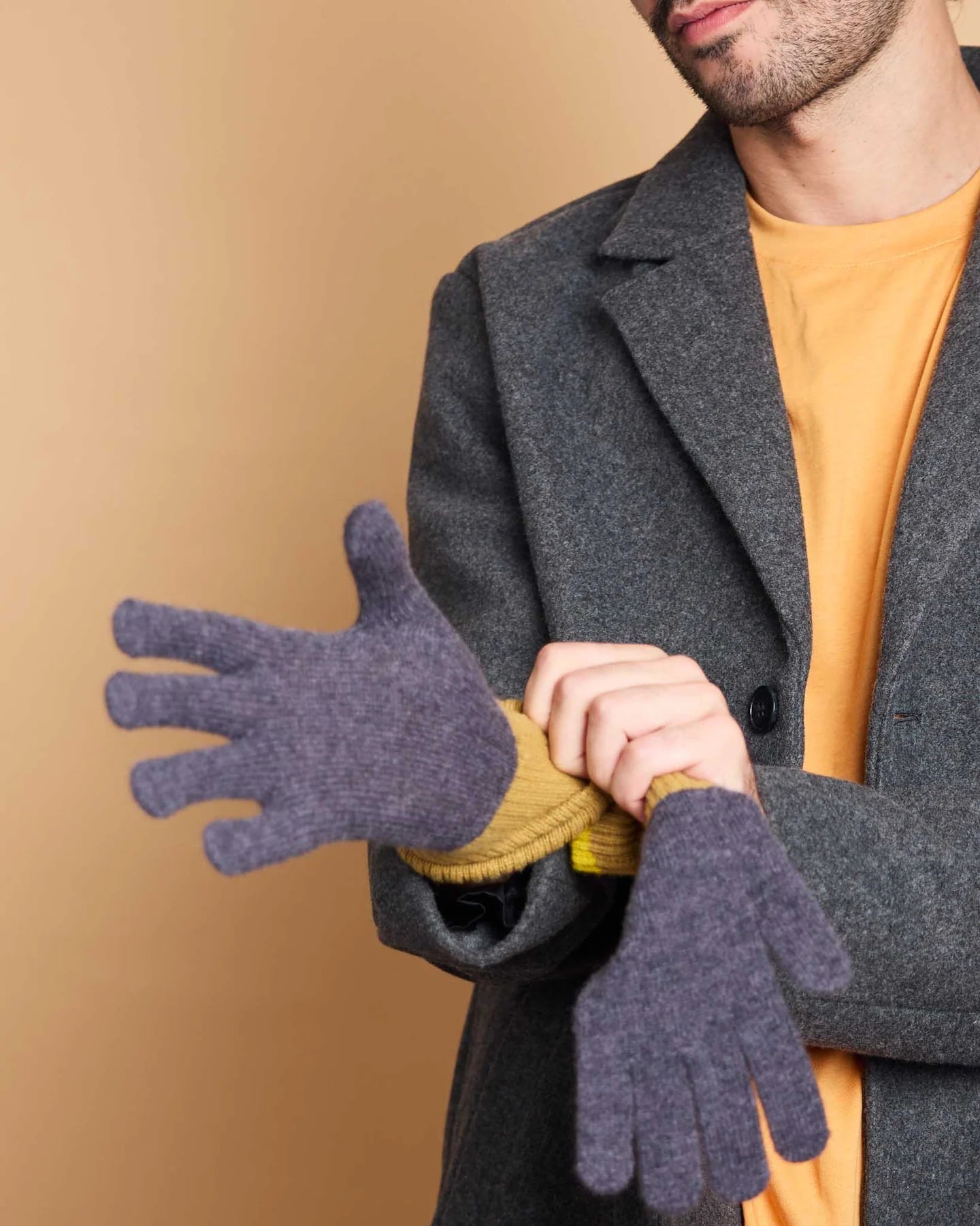A man wearing a dark gray wool coat over a mustard yellow t-shirt, adjusting a pair of dark purple knit gloves.