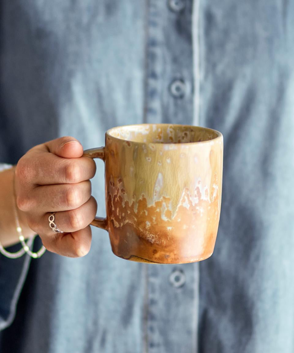Hand holding a brown ceramic mug with a blurred gray shirt in the background