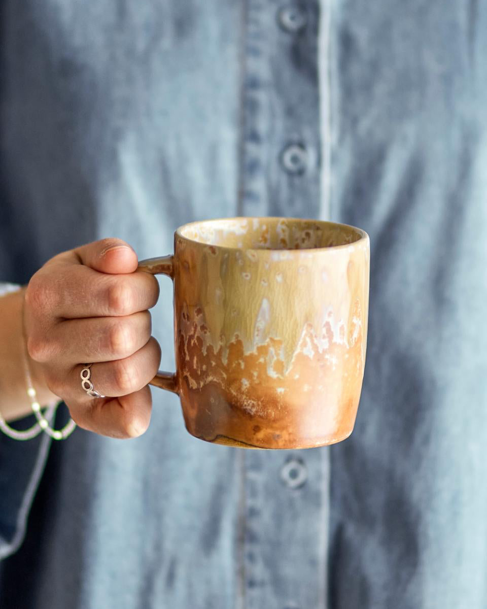 Hand holding a brown ceramic mug with a blurred gray shirt in the background