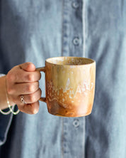 Hand holding a brown ceramic mug with a blurred gray shirt in the background