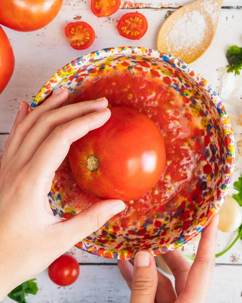 A person holding a multicolored ceramic grater plate with a floral and geometric pattern, over a background that includes tomatoes, garlic, and parsley.