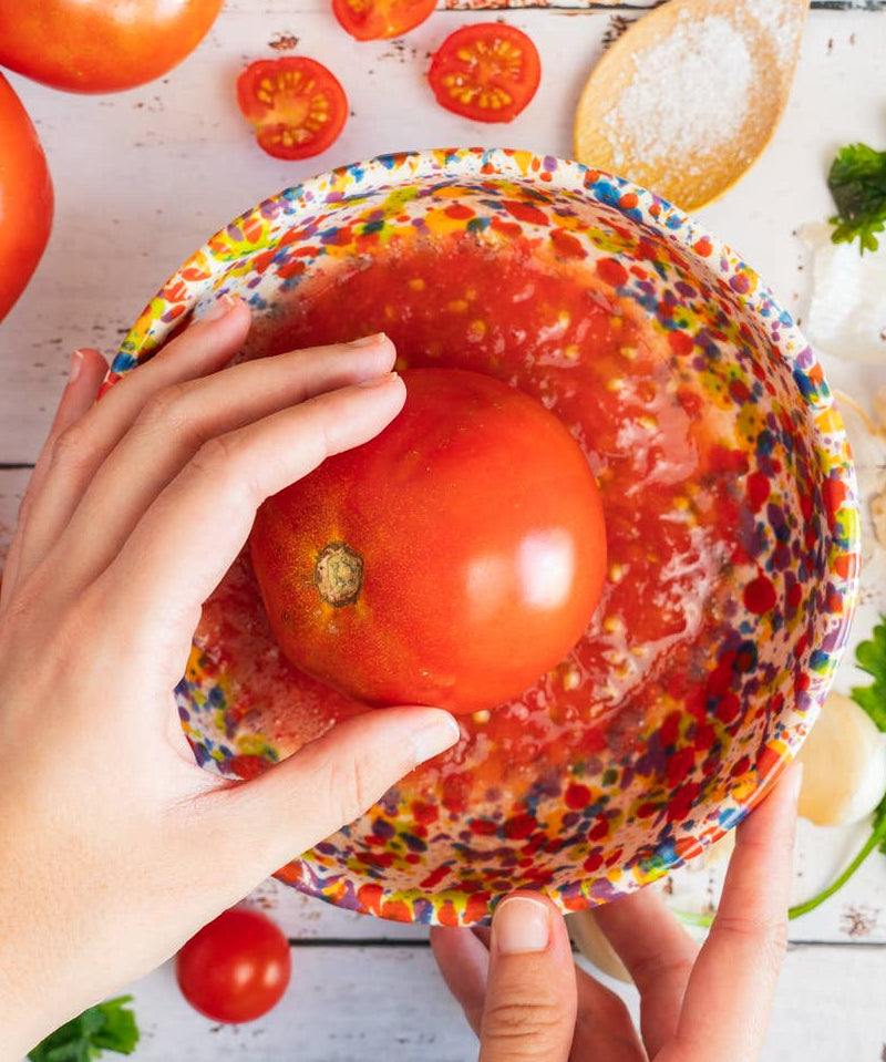 A person holding a multicolored ceramic grater plate with a floral and geometric pattern, over a background that includes tomatoes, garlic, and parsley.