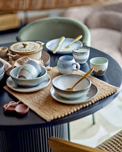 Ceramic dishes on a round table with woven placemats and wooden spoons.