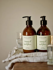 Two dark brown glass bottles, black pump dispensers, white labels with sage green writing, seaweed and samphire shampoo and conditioner by Plum and Ashby