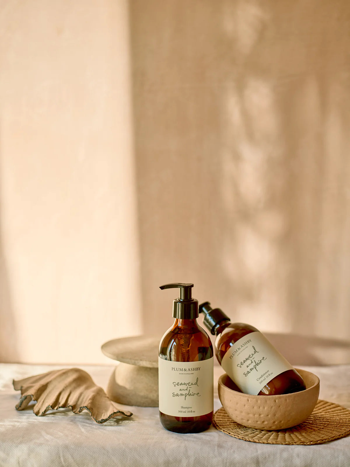Two dark brown glass bottles, black pump dispensers, white labels with sage green writing, seaweed and samphire shampoo and conditioner by Plum and Ashby
