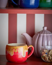 Red mug with gold accents on a shelf with a striped wall in the background