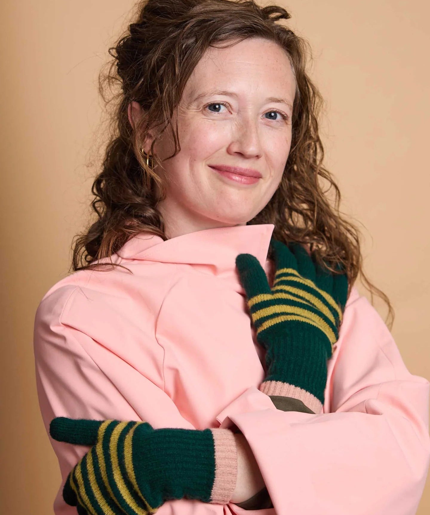 A woman with curly brown hair wearing a pink jacket and green knit gloves with yellow stripes, posing against a solid beige background.