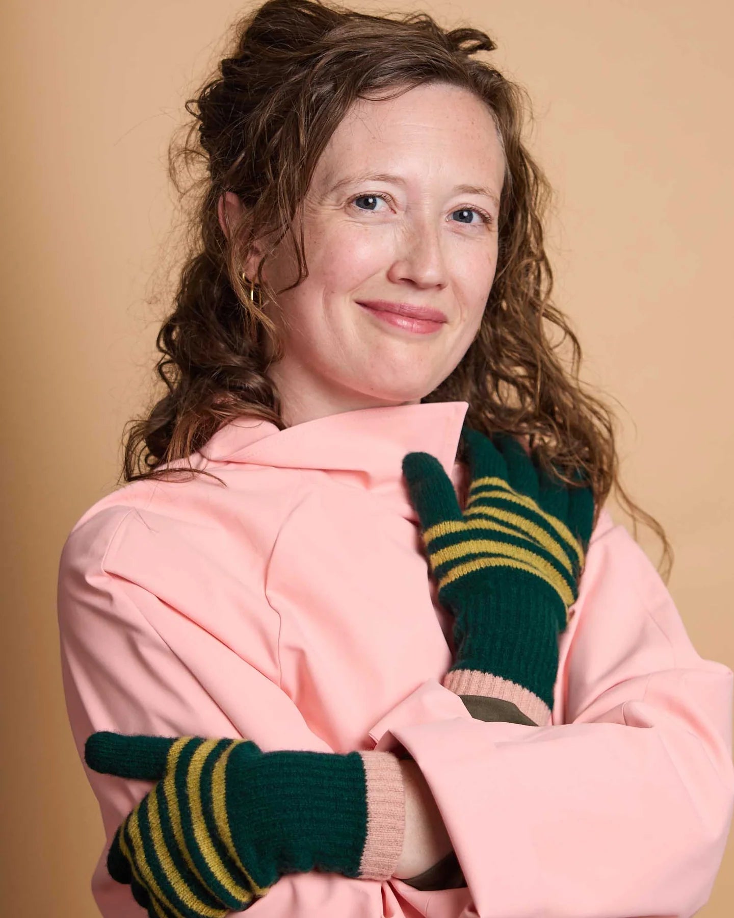 A woman with curly brown hair wearing a pink jacket and green knit gloves with yellow stripes, posing against a solid beige background.