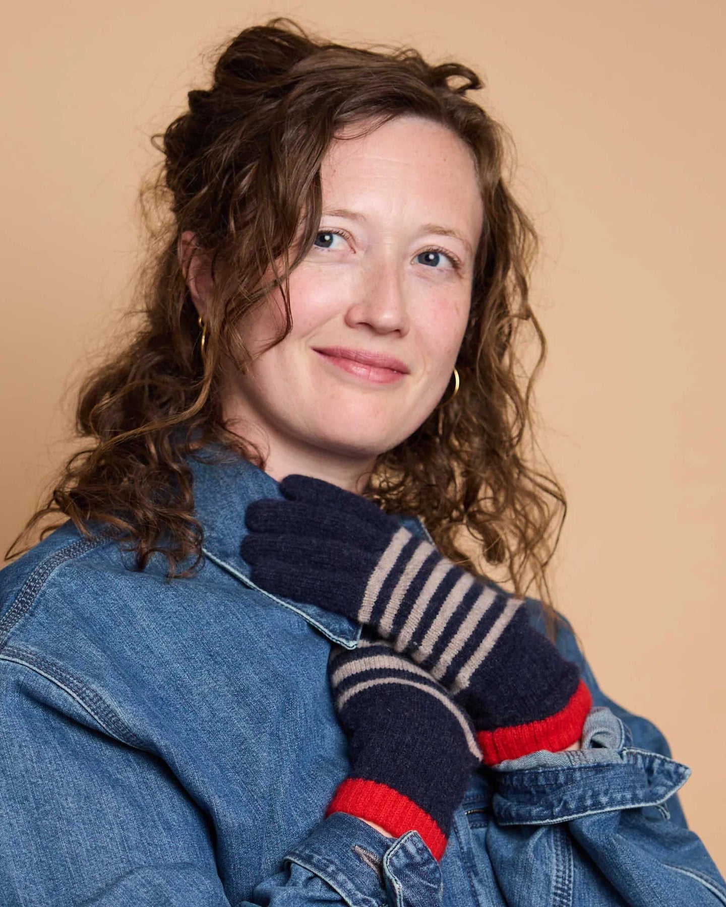 A woman with curly brown hair wearing a blue denim jacket and navy blue knit gloves with beige stripes and red cuffs, posing against a solid beige background.