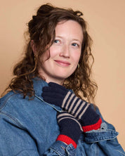 A woman with curly brown hair wearing a blue denim jacket and navy blue knit gloves with beige stripes and red cuffs, posing against a solid beige background.