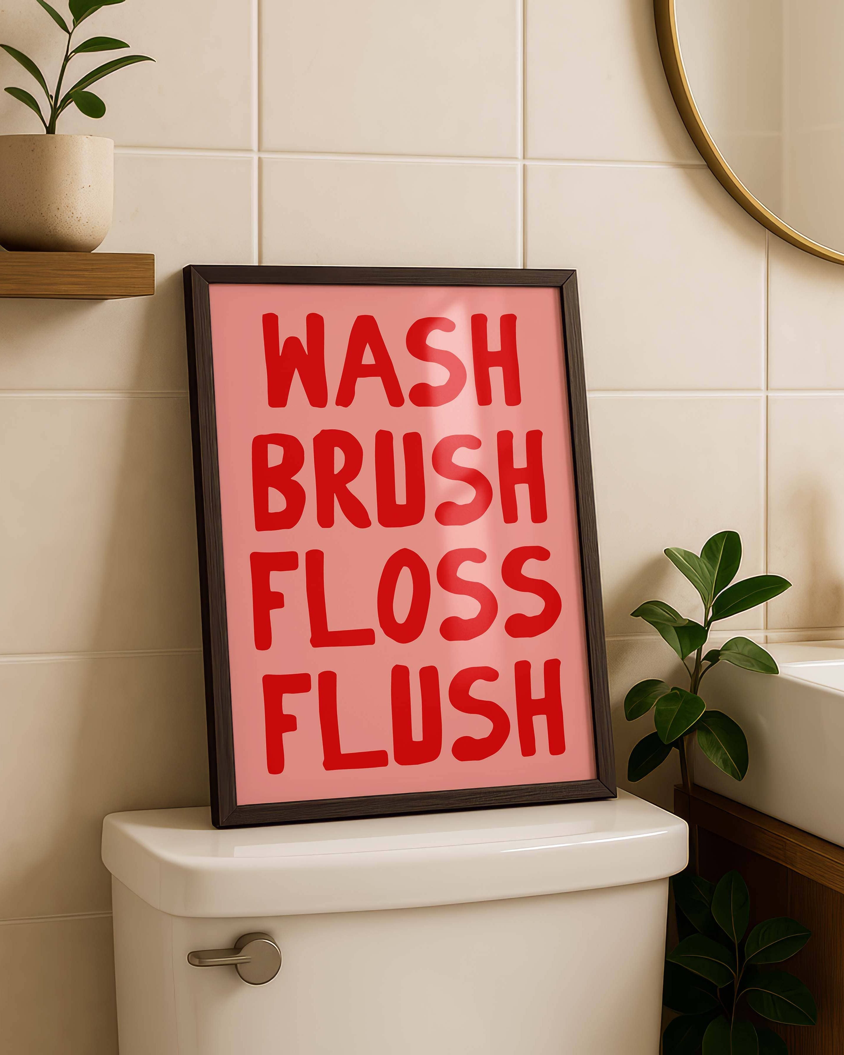 A framed wall art print with the words 'WASH BRUSH FLOSS FLUSH' in red lettering on a pink background, leaning on a white toilet in a bathroom setting.