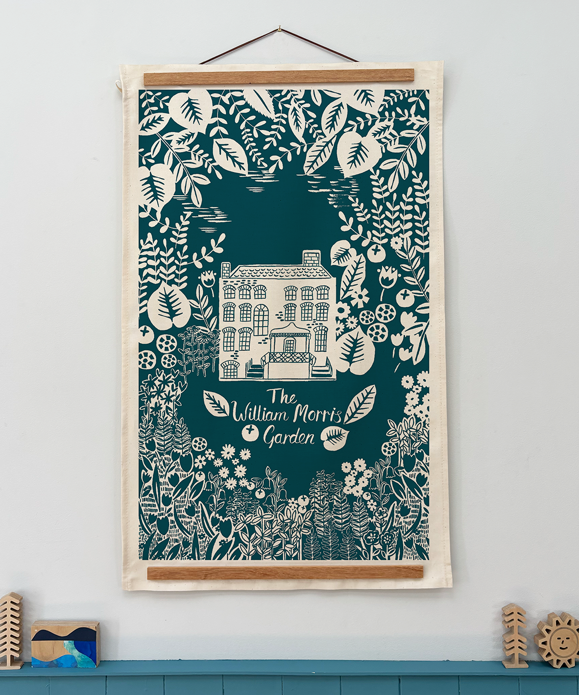 Decorative tea towel with a house design and floral elements on a teal background, framed and hanging on a wall.